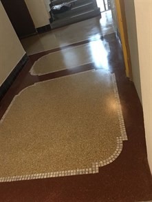 Wypolerowana podłoga z lastryko w korytarzu, z dekoracyjnymi wstawkami w kształcie łezek obramowanymi mozaiką. Widoczne schody w tle.