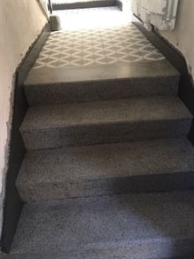 Betonowe schody z szarymi stopniami i wzorzystą posadzką na podeście, widok z dołu w kierunku jasnego wyjścia.