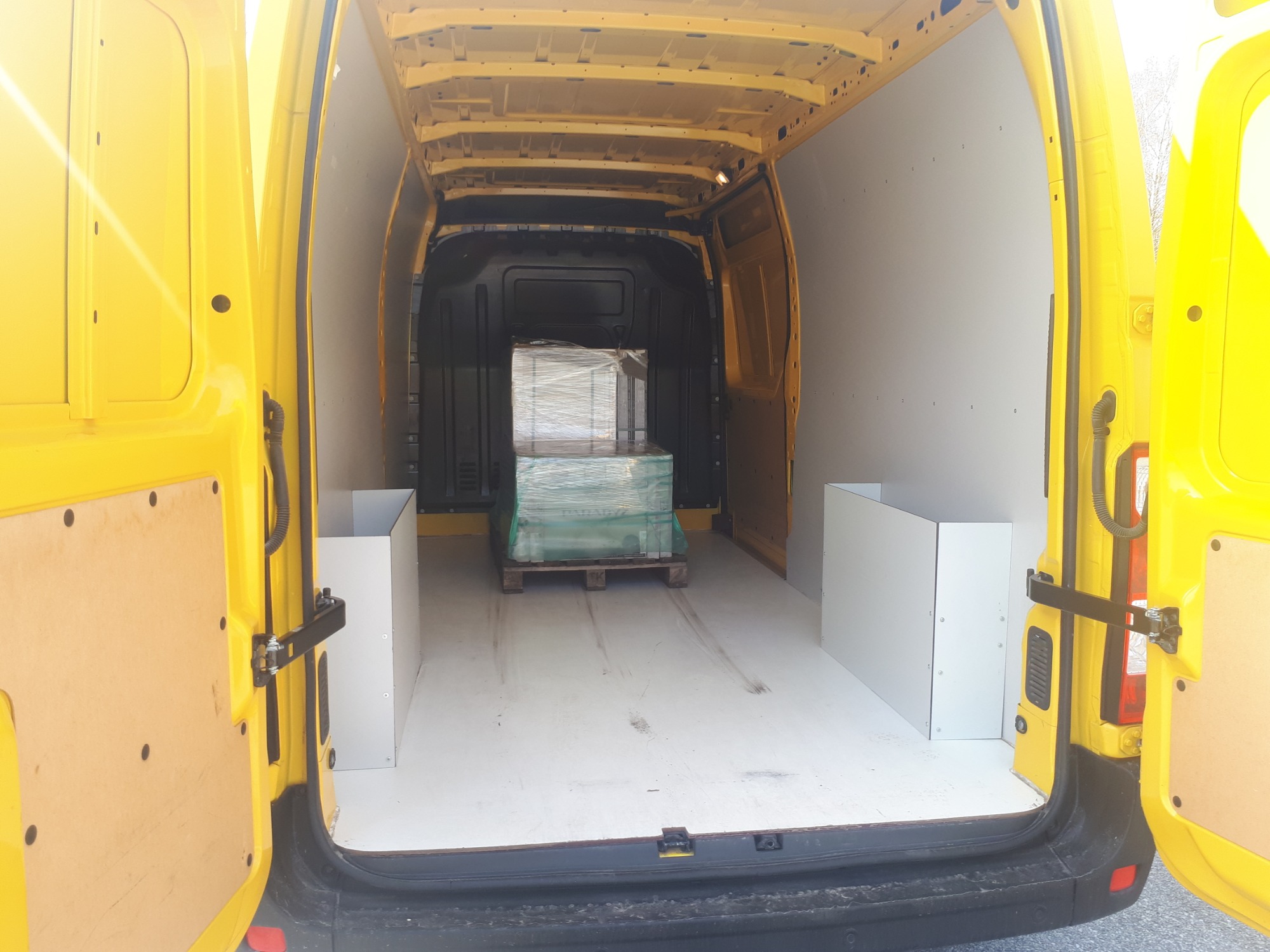 Wnętrze żółtego samochodu dostawczego z otwartymi drzwiami, zabezpieczone białą zabudową, z paletą owiniętą folią na środku, widoczne ślady użytkowania na podłodze.