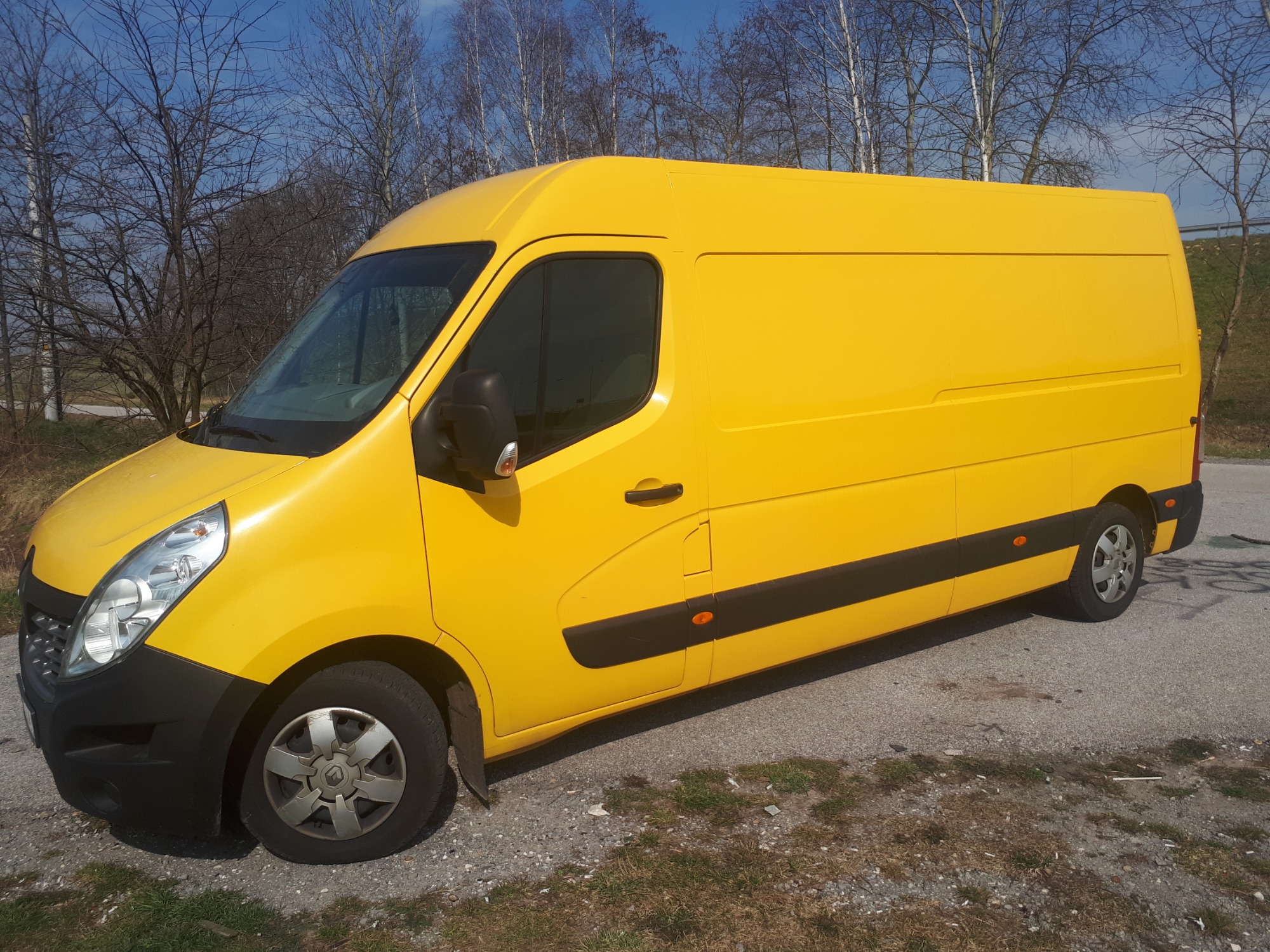 Żółty samochód dostawczy Renault Master zaparkowany na poboczu drogi, z widocznymi drzewami bez liści w tle i fragmentem niebieskiego nieba.