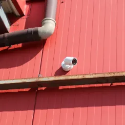 Biała kamera monitoringu zamontowana na czerwonej ścianie blaszanej budynku, widoczna rura spustowa i skorodowany metalowy profil.