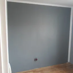 Świeżo pomalowana szara ściana z białą listwą przypodłogową i drewnianą podłogą, widoczna puszka elektryczna.