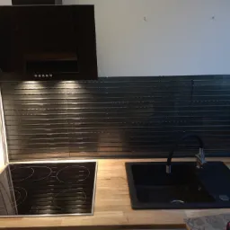 Nowoczesna kuchnia z czarną płytą indukcyjną, czarnym zlewem i błyszczącymi, poziomymi płytkami nad blatem, oświetlona lampkami podszafkowymi.