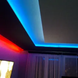 Nowoczesny sufit z podwieszanymi elementami i oświetleniem LED w kolorach czerwonym i niebieskim, widoczne okno z zasłonami i fragment telewizora.