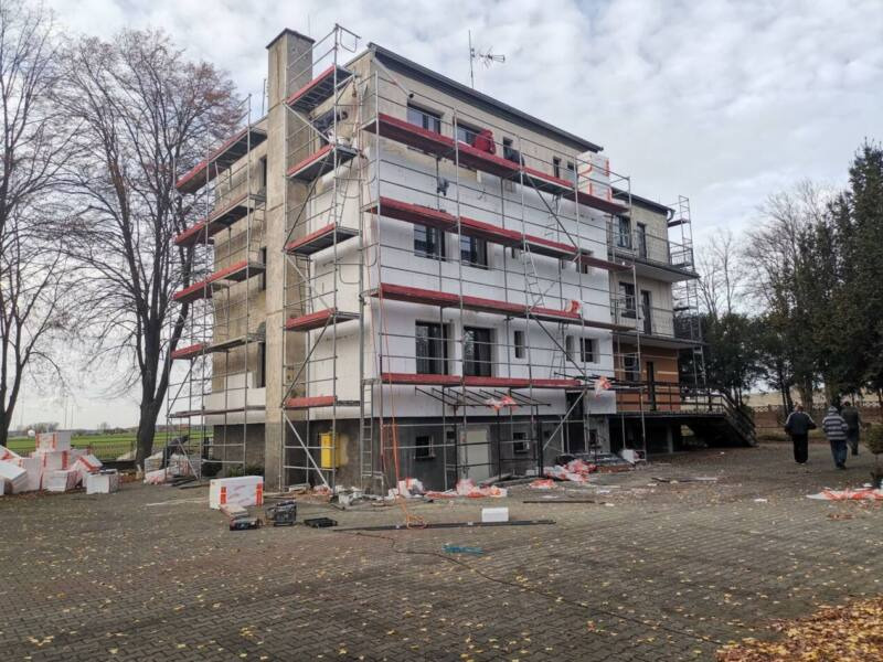 Trzy kondygnacyjny budynek w trakcie prac, częściowo ocieplony styropianem, z rozstawionymi rusztowaniami, widoczne materiały budowlane i narzędzia na brukowanym placu przed budynkiem, pochmurne...