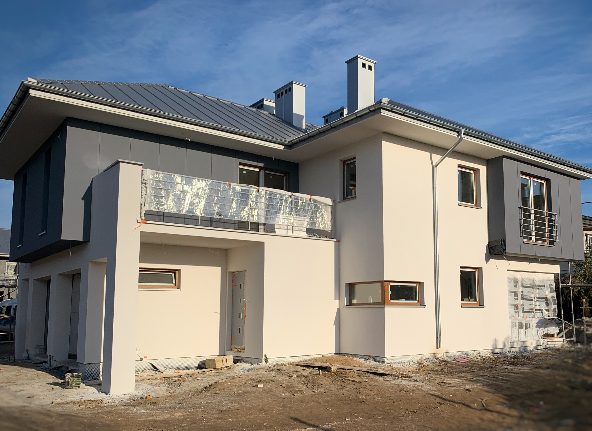 Nowoczesny dom jednorodzinny w trakcie prac elewacyjnych, z widocznym rusztowaniem i folią ochronną na balkonie, świeżo otynkowane ściany w kolorze kremowym i szarym, teren wokół domu w trakcie...