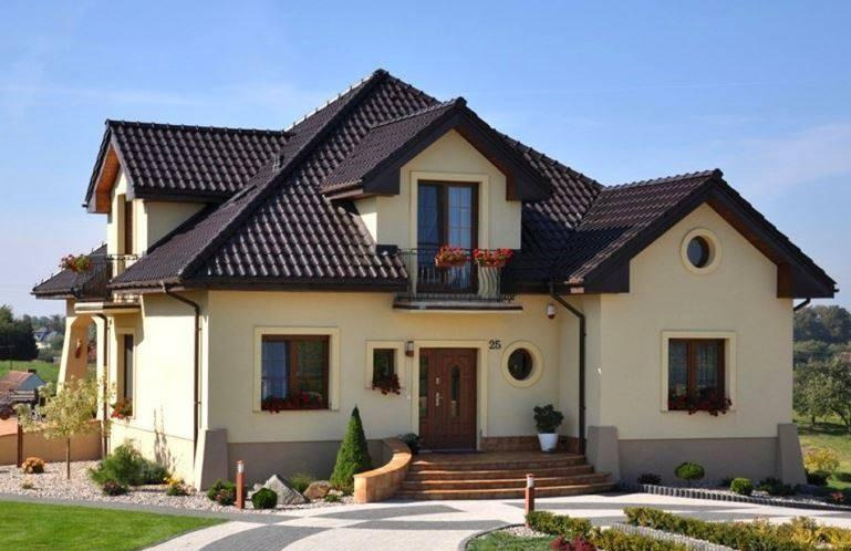 Jasny, dwukondygnacyjny dom jednorodzinny z ciemnobrązowym dachem, balkonem z kwiatami i numerem domu '25', otoczony zadbanym ogrodem z kostki brukowej i niską roślinnością.