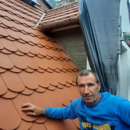 Mężczyzna w niebieskiej bluzie prezentuje ułożoną dachówkę karpiówkę w kolorze ceglastym, z widocznym fragmentem rusztowania i czarną folią ochronną na elewacji.