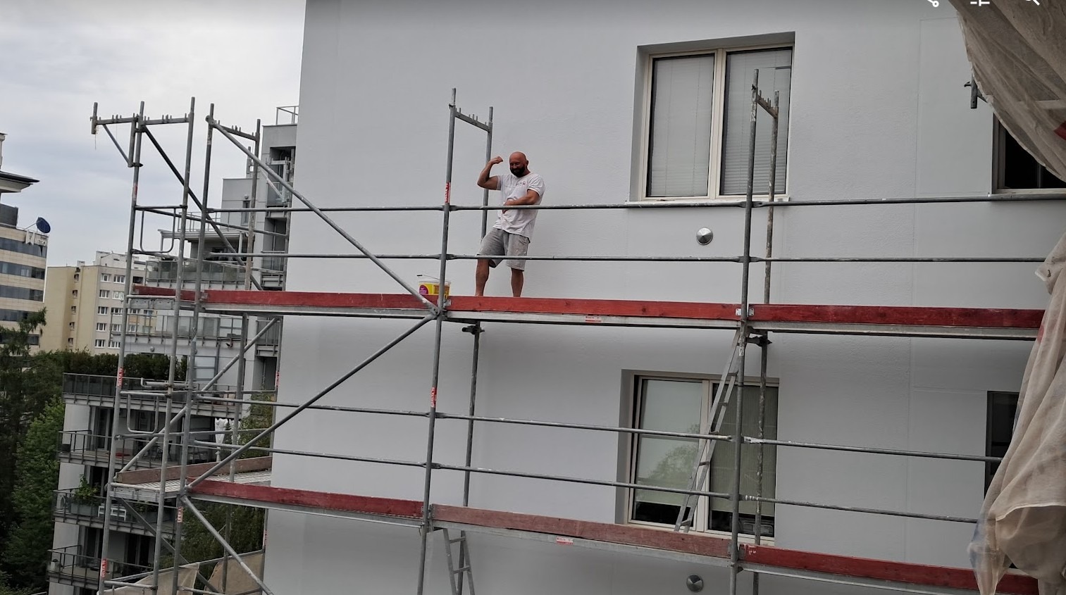 Mężczyzna na rusztowaniu prezentuje biceps po pomalowaniu fragmentu białej elewacji budynku wielorodzinnego; wiadro z farbą obok, widok na sąsiednie budynki w tle.