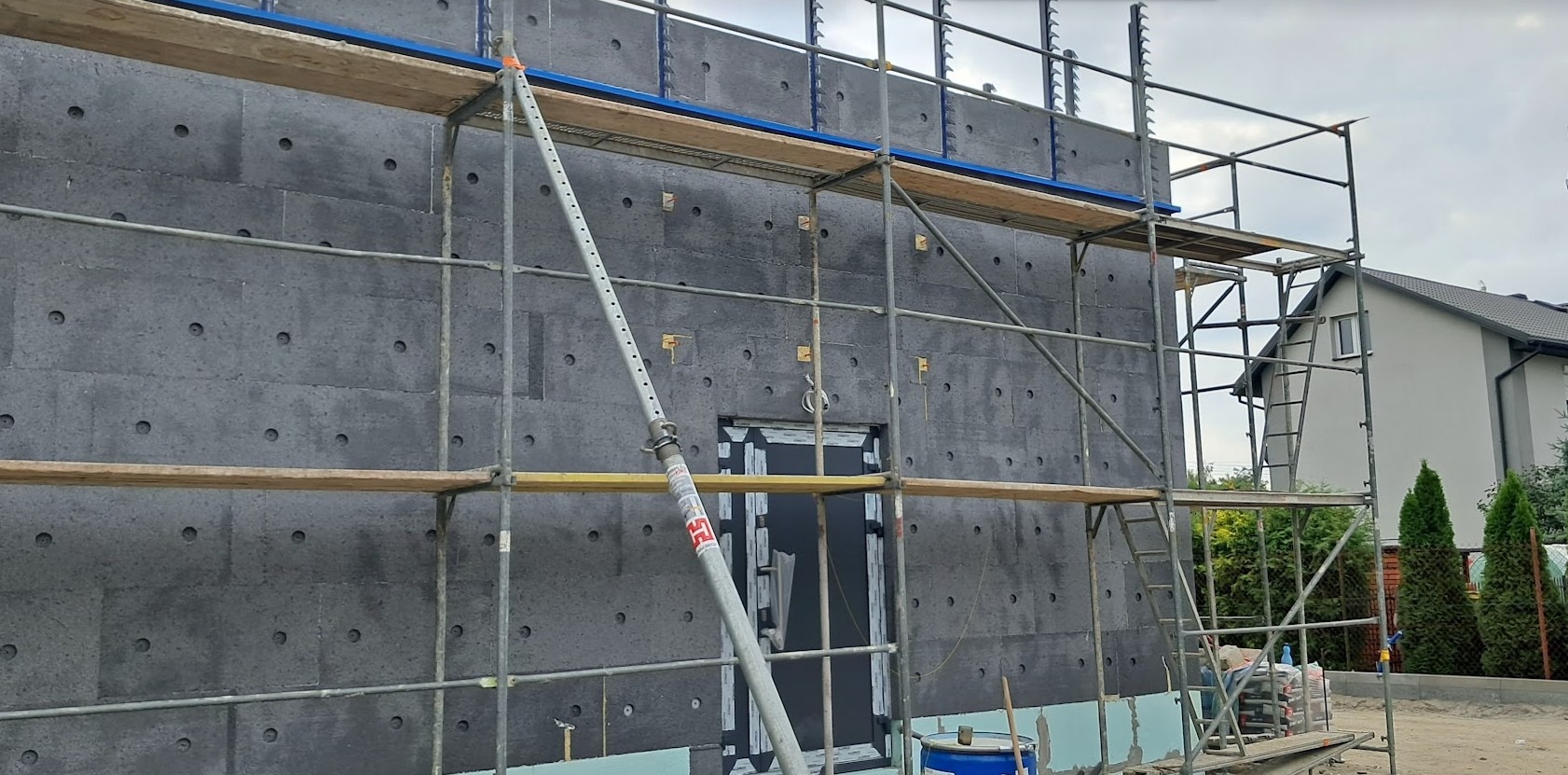 Ściana budynku w Wołominie pokryta szarymi płytami izolacyjnymi z widocznymi otworami, rusztowanie metalowe przy ścianie, okno tymczasowo zasłonięte, przygotowania do dalszych prac.