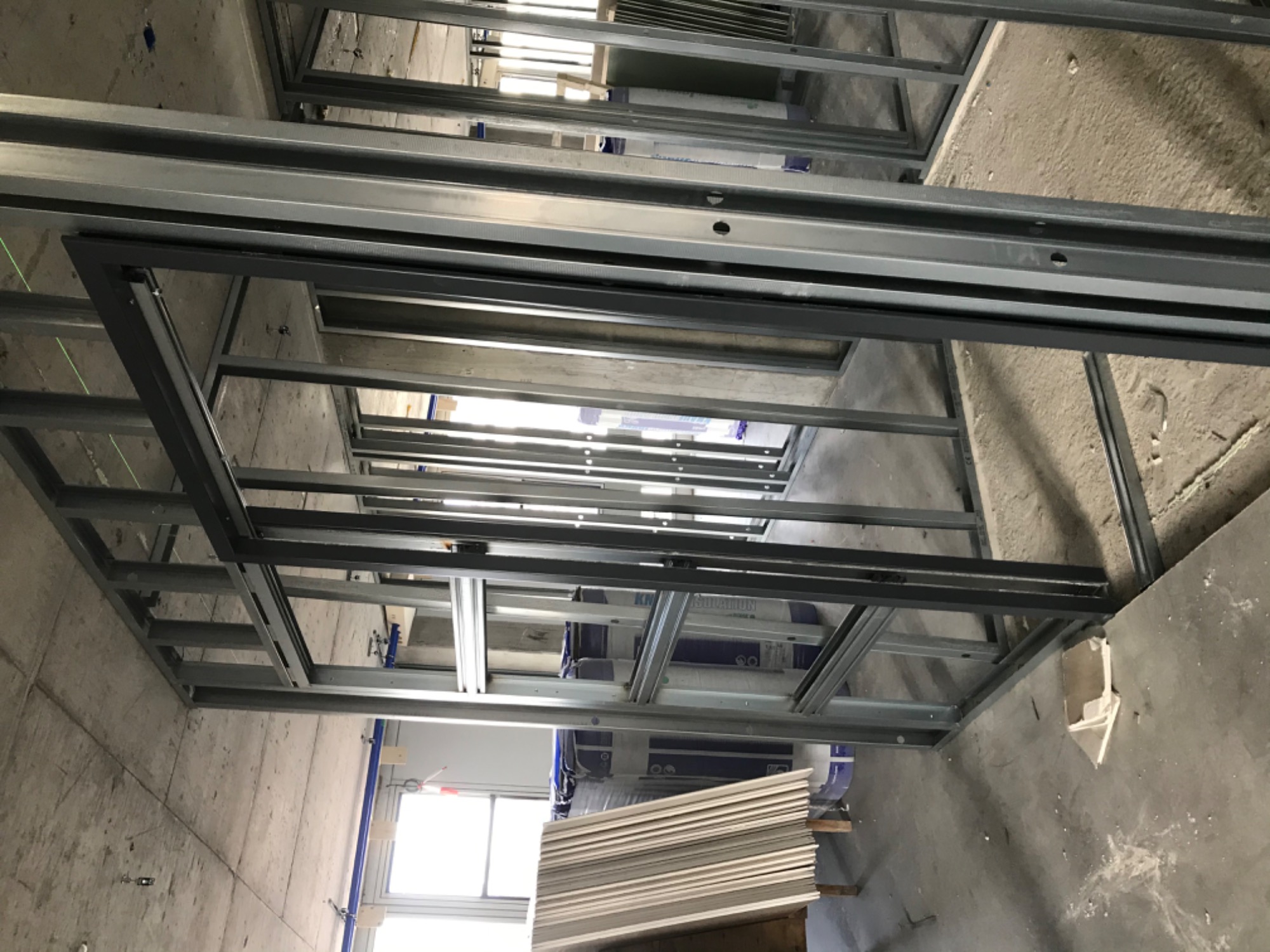 Metalowa konstrukcja sufitu podwieszanego w trakcie montażu, widoczne płyty gotowe do instalacji, surowy betonowy strop w tle.