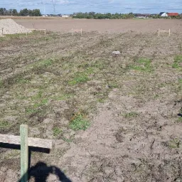 Wytyczony teren pod budowę z wbitymi, pomalowanymi na zielono słupkami i poprzeczkami, w tle hałdy ziemi i pole uprawne, widoczne słupy wysokiego napięcia.