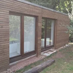 Nowoczesny dom z elewacją z drewnianych desek, widoczne okna z brązowymi ramami, trawnik i drzewa w tle.