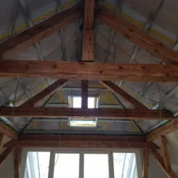 Poddasze z widoczną konstrukcją drewnianą, ociepleniem z folii i wełny mineralnej, okno dachowe w centralnej części dachu, widok od dołu.