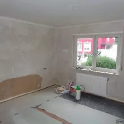 Pokój w trakcie remontu: ściany z widocznymi śladami po naprawach, surowy tynk, okno z grzejnikiem, materiały budowlane i narzędzia na podłodze, listwa dekoracyjna pod sufitem.