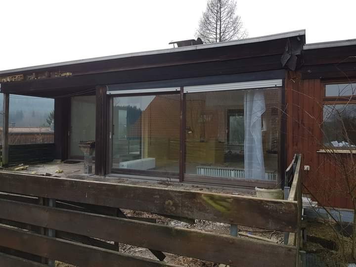 Drewniana weranda z przesuwnymi oknami, widoczne ślady remontu, stare drewniane balustrady, częściowo zniszczony dach.