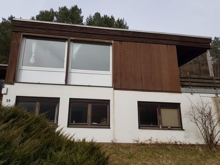 Dwukondygnacyjny dom z białymi ścianami i drewnianymi elementami, widoczne okna z zasłonami i roletami, numer 39 na ścianie, otoczenie z zielenią.