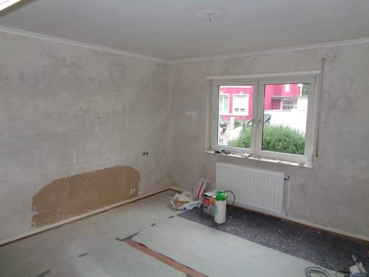 Pokój w trakcie remontu: ściany z widocznymi śladami po naprawach, surowy tynk, okno z grzejnikiem, materiały budowlane i narzędzia na podłodze, listwa dekoracyjna pod sufitem.