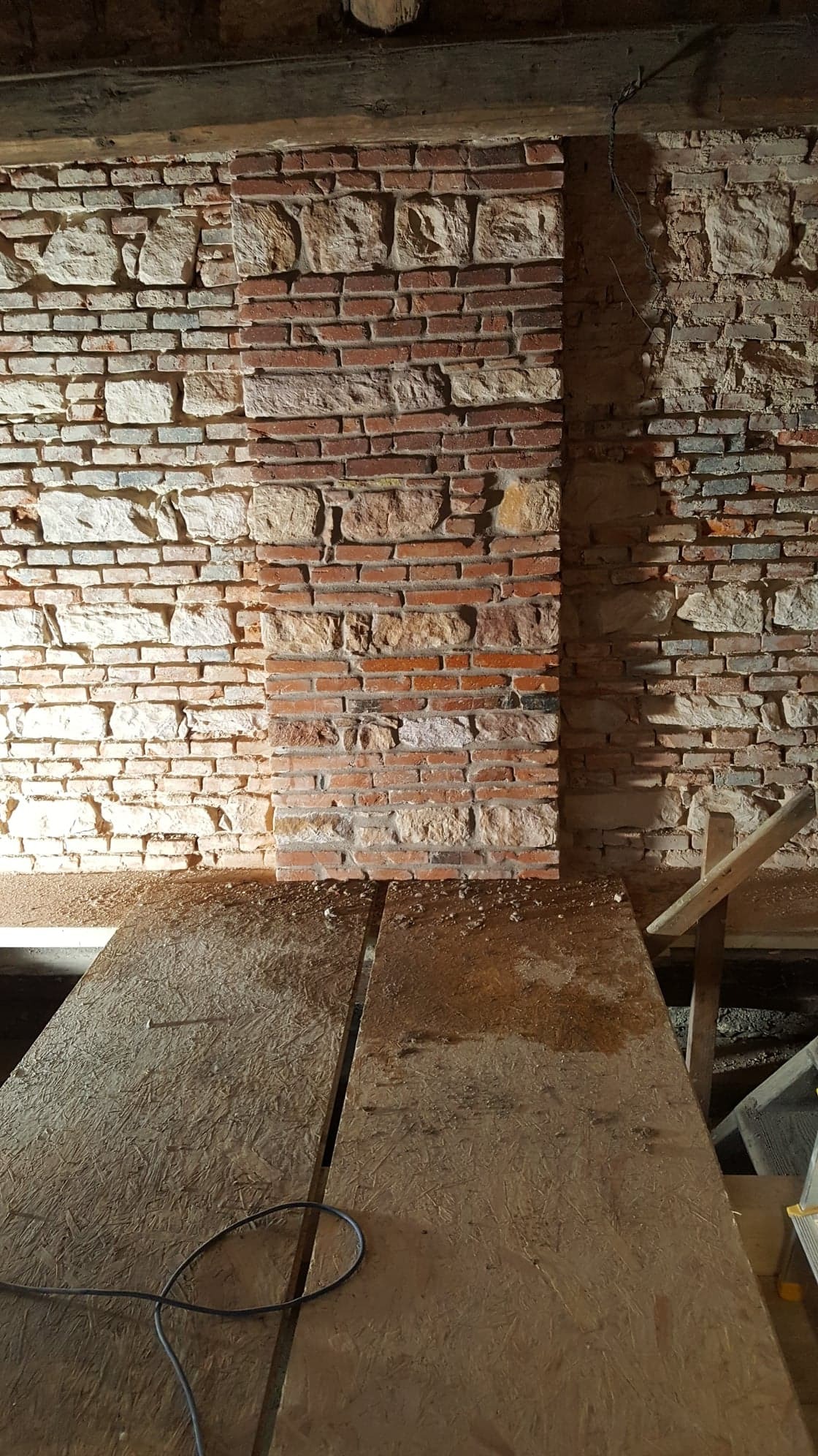 Fragment murowanej ściany z cegły i kamienia, z widocznymi śladami prac remontowych i deskami na podłodze.