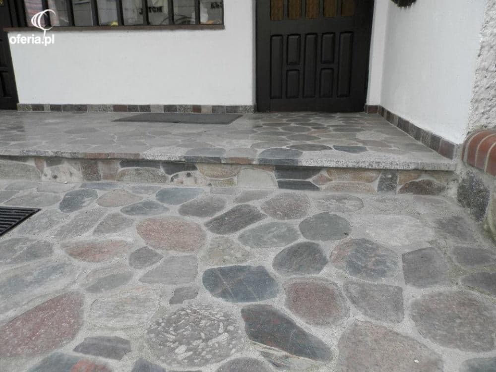 Wykończony kamienny podjazd przed wejściem do domu, z ciemnymi drzwiami i fragmentem okna w tle.