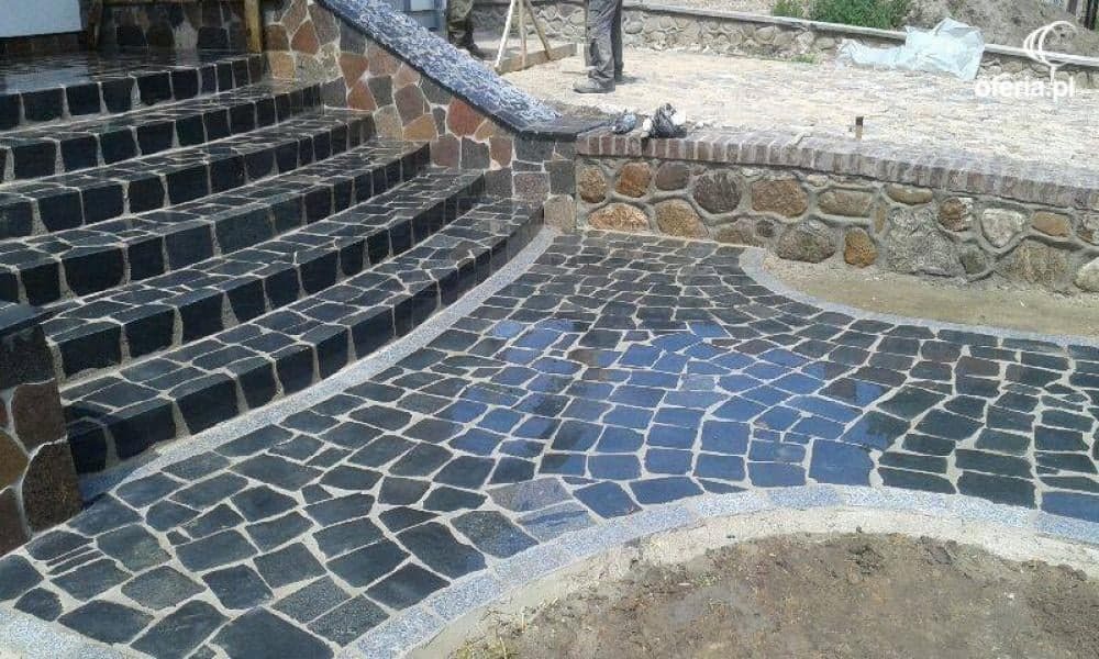 Schody zewnętrzne i ścieżka wyłożone czarną kostką granitową, z obrzeżem z jasnego kamienia, prowadzące do kamiennego murku.