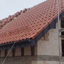 Montaż czerwonej dachówki ceramicznej na nowym dachu z szarą rynną, widoczne zabezpieczenia dla dekarzy i rusztowanie przy ścianie budynku w trakcie budowy.