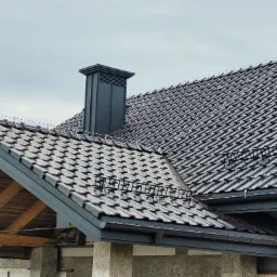 Nowy dach z ciemnej dachówki ceramicznej, widoczne rynny, komin i zabezpieczenie przeciwśniegowe. Drewniana konstrukcja dachu w trakcie budowy.