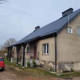 CM Dachy - Dom jednorodzinny z nowym, grafitowym dachem z blachodachówki, widoczny z perspektywy ogrodu. Czerwony samochód zaparkowany obok. Nowe rynny w kolorze dachu.