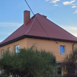 CM Dachy - Dom z nowym, czerwonym dachem i kominem z cegły, widoczny z poziomu ogrodu. Rynny w kolorze grafitowym. Błękitne niebo w tle.