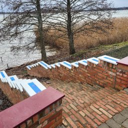 CM Dachy - Mur z cegły klinkierowej z białymi i niebieskimi akcentami, tworzący schody w dół w kierunku jeziora. Widoczne drzewa i trzciny w tle, pochmurne niebo.