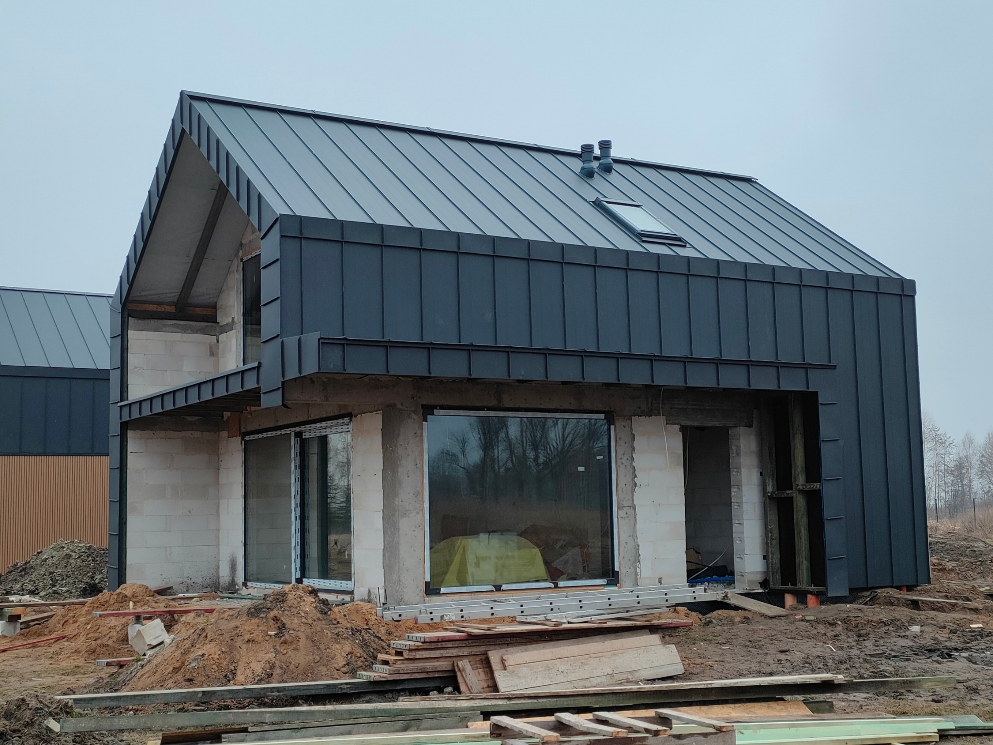 Dom w trakcie budowy z grafitowym dachem blaszanym na rąbek stojący, widoczne okna dachowe i kominy wentylacyjne, ściany z pustaków.
