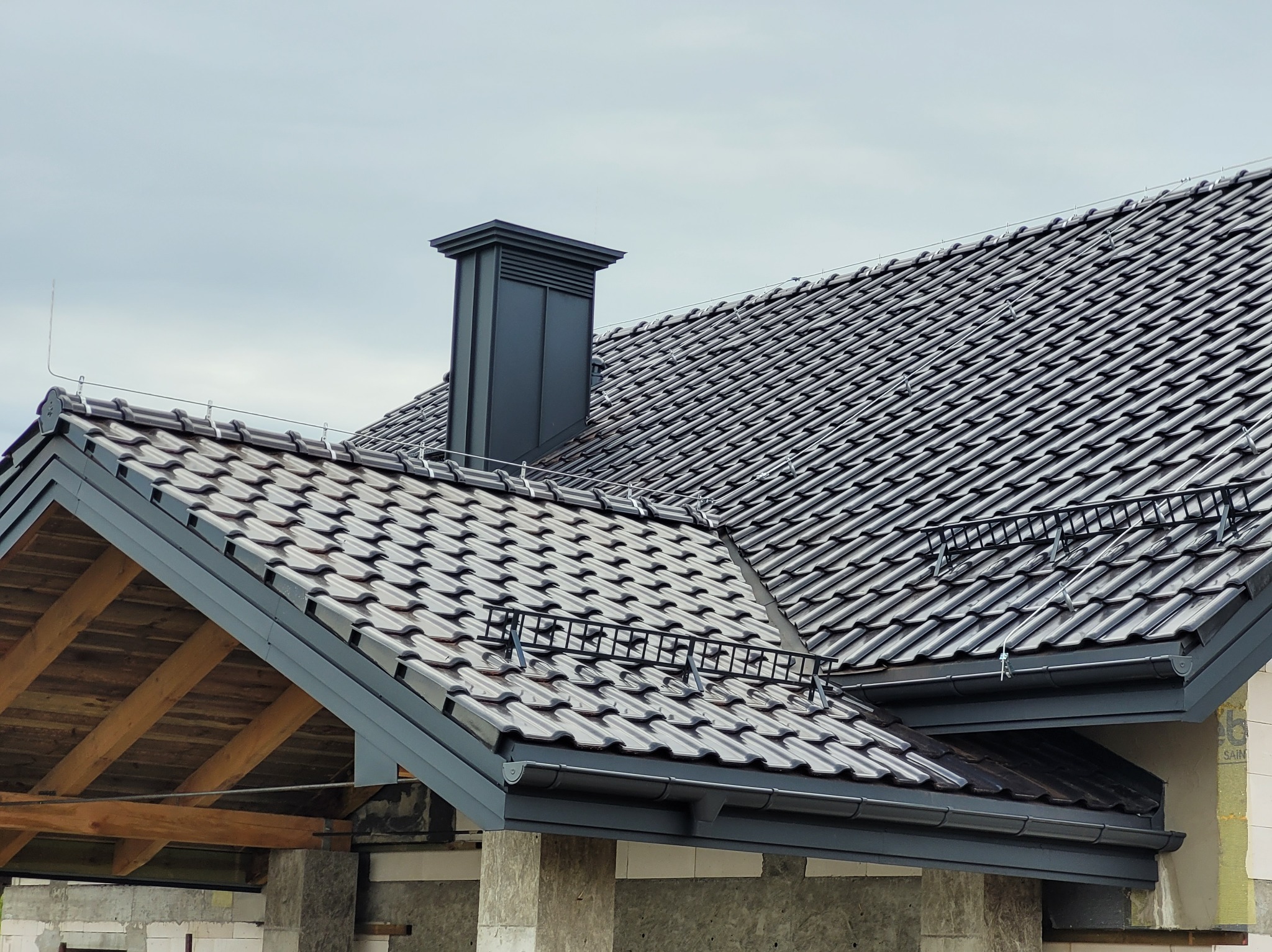 Nowy dach z ciemnej dachówki ceramicznej, widoczne rynny, komin i zabezpieczenie przeciwśniegowe. Drewniana konstrukcja dachu w trakcie budowy.