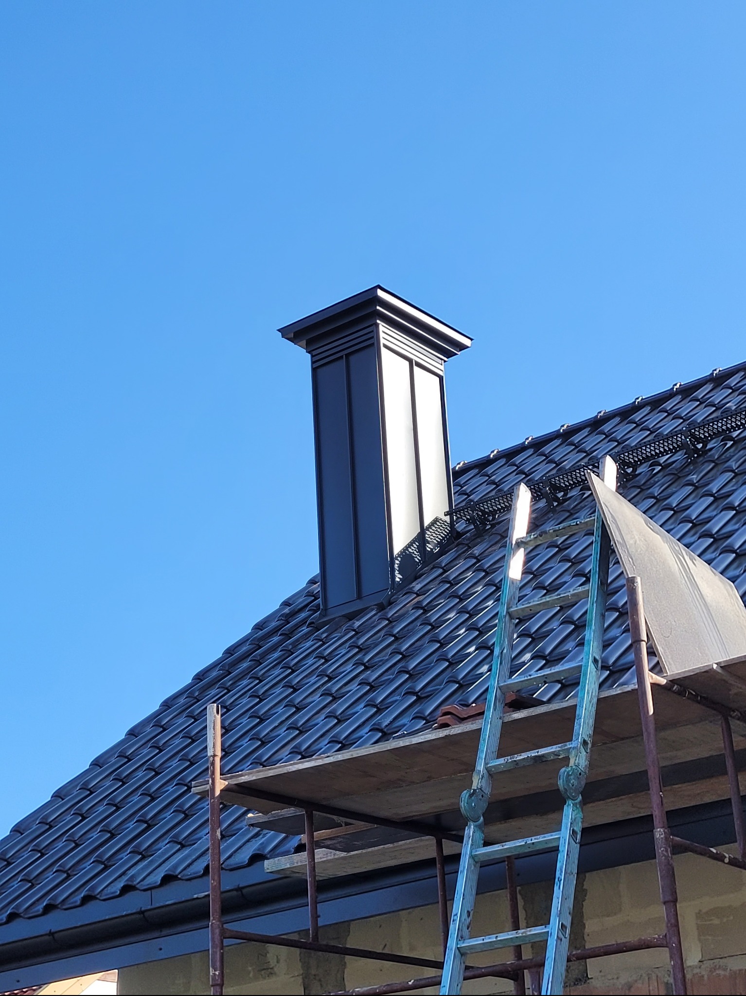 Czarny komin z blachy na dachu z ciemnej dachówki ceramicznej, widoczne rusztowanie i drabina. Błękitne niebo w tle.