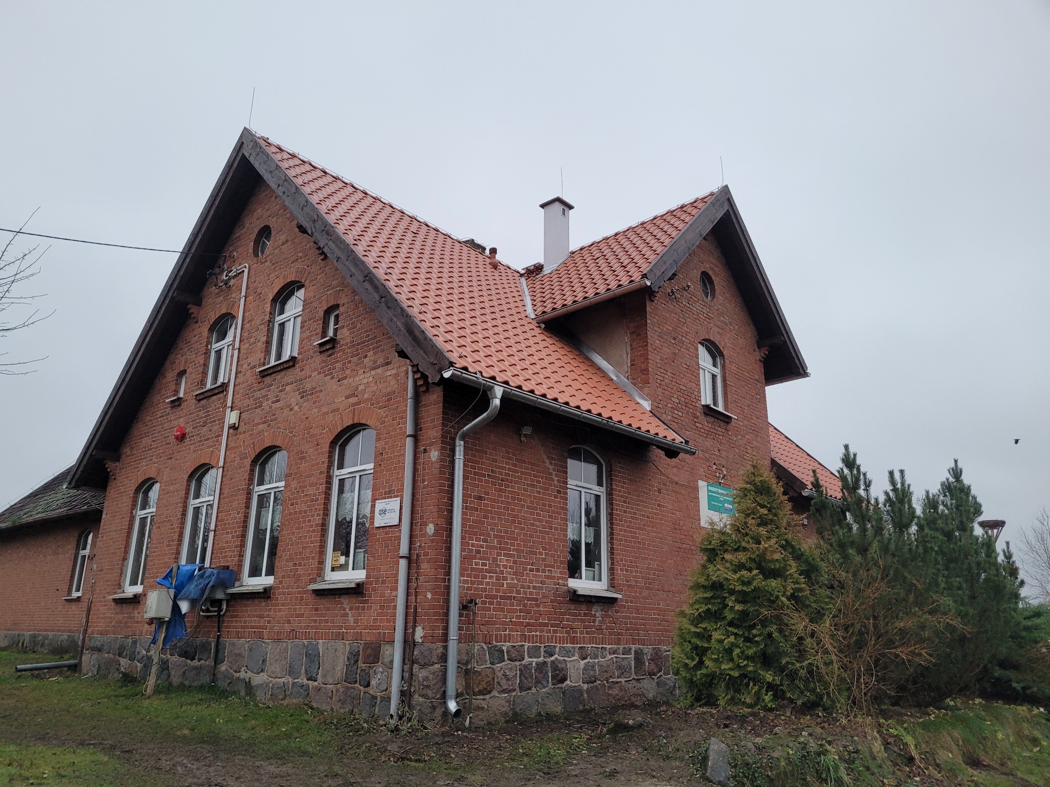Ceglany budynek z czerwonym dachem ceramicznym, widoczne rynny i komin. Uwagę zwraca nietypowa architektura i połączenie cegły z kamienną podmurówką.