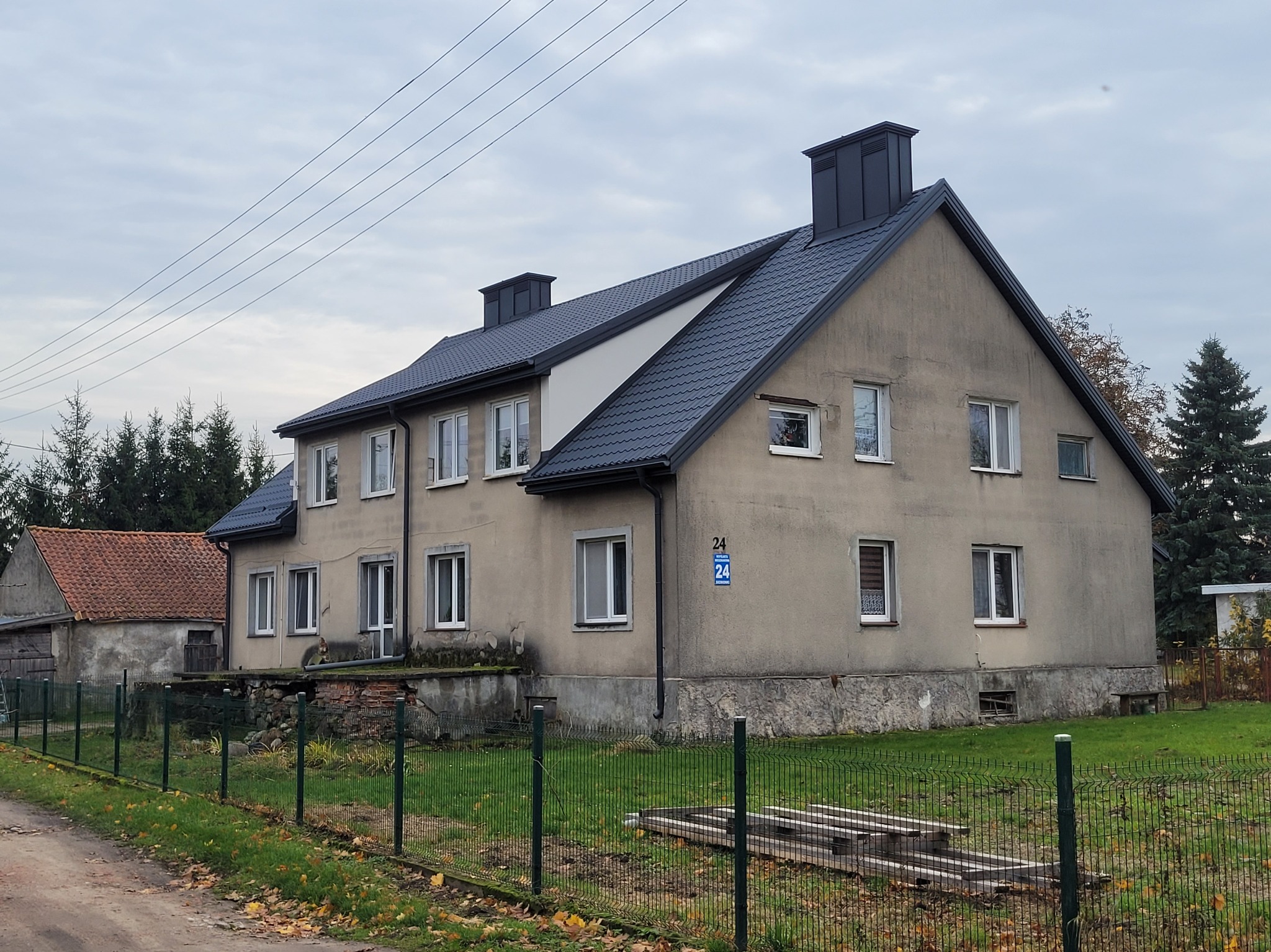 Dwukondygnacyjny budynek z ciemnoszarym dachem z blachy trapezowej i kominami w tym samym kolorze. Widoczny numer domu '24'.