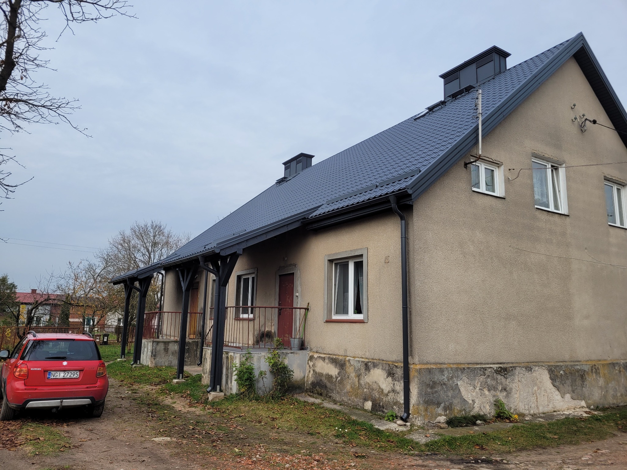 Dom jednorodzinny z nowym, grafitowym dachem z blachodachówki, widoczny z perspektywy ogrodu. Czerwony samochód zaparkowany obok. Nowe rynny w kolorze dachu.