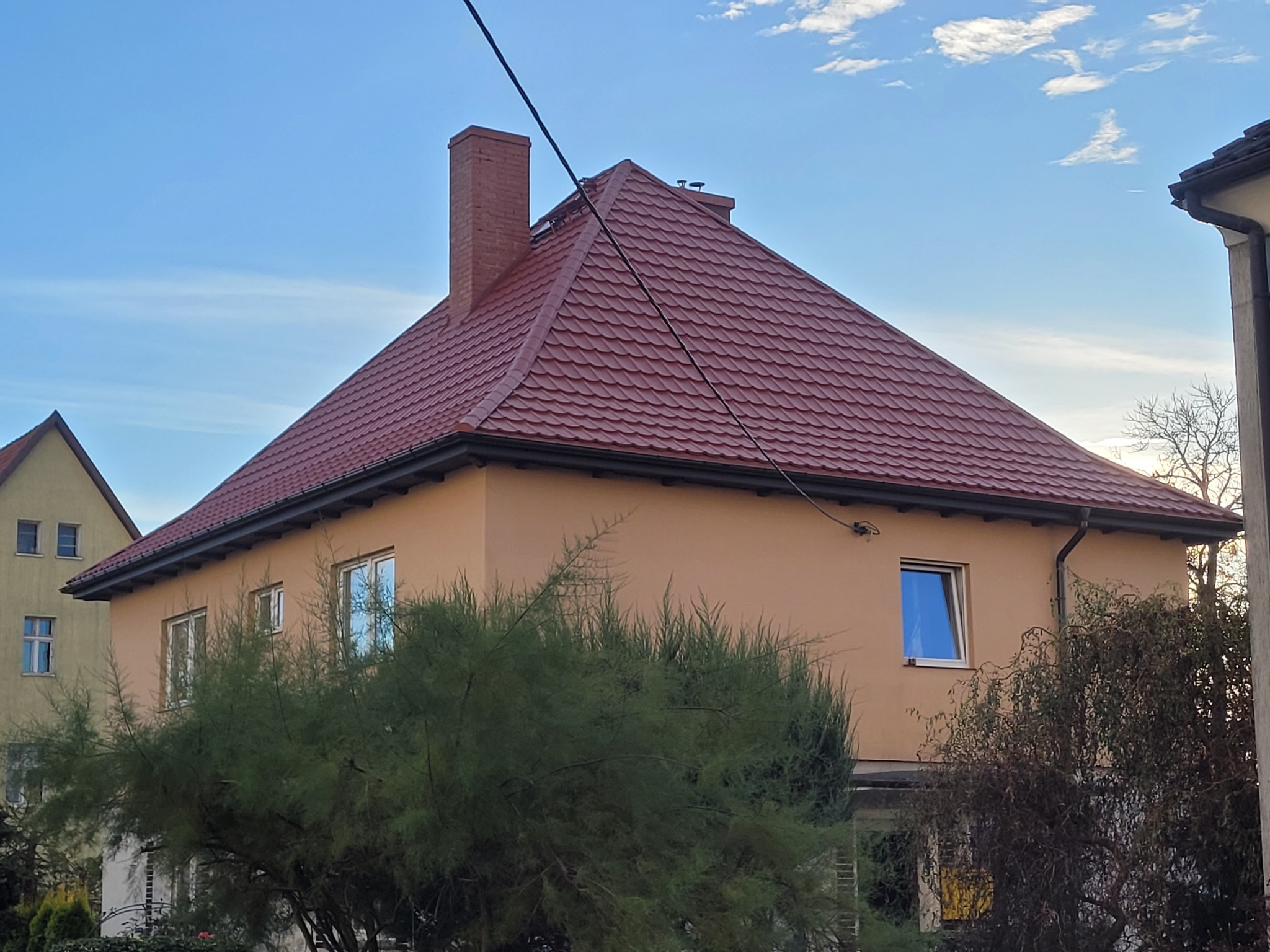 Dom z nowym, czerwonym dachem i kominem z cegły, widoczny z poziomu ogrodu. Rynny w kolorze grafitowym. Błękitne niebo w tle.