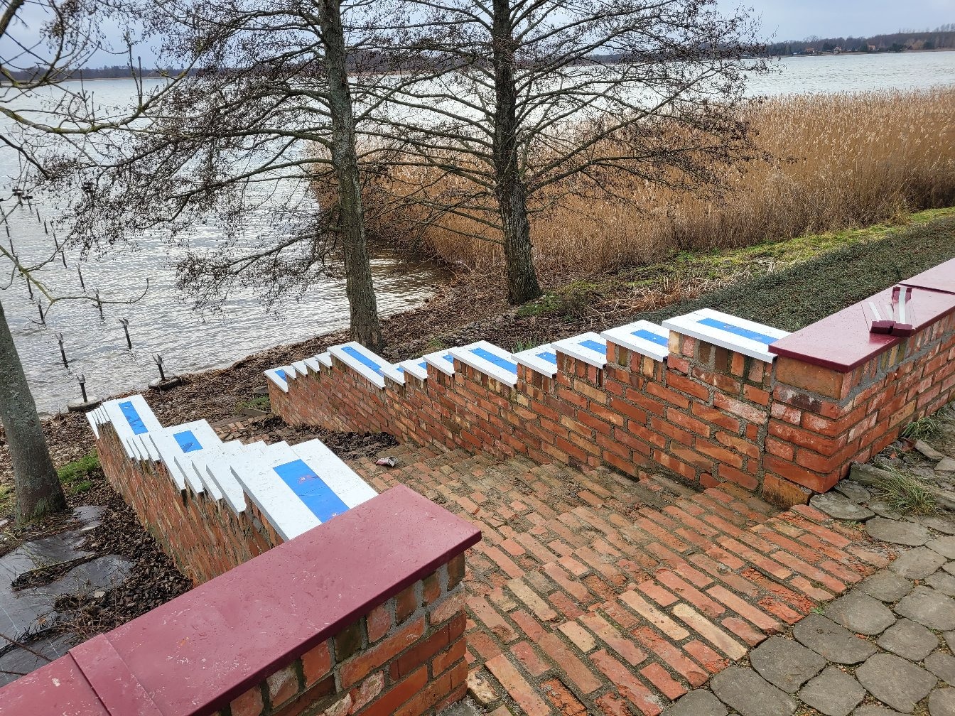Mur z cegły klinkierowej z białymi i niebieskimi akcentami, tworzący schody w dół w kierunku jeziora. Widoczne drzewa i trzciny w tle, pochmurne niebo.
