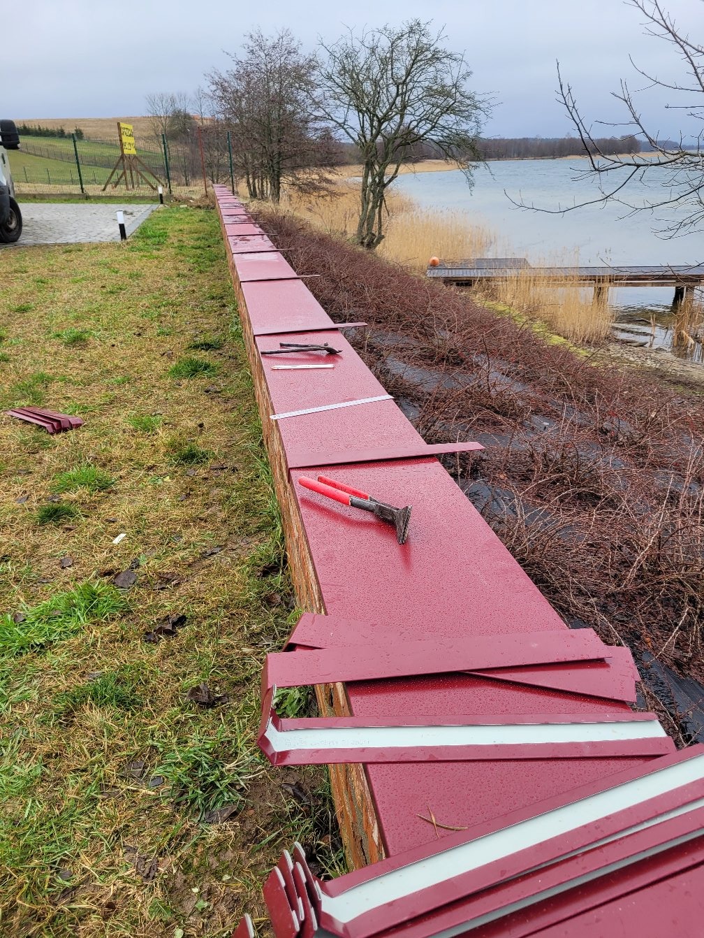 Czerwone, metalowe elementy obróbki blacharskiej ułożone wzdłuż krawędzi dachu, narzędzie dekarskie na wierzchu, w tle jezioro i drzewa. Widok z bliska, pochmurny dzień.