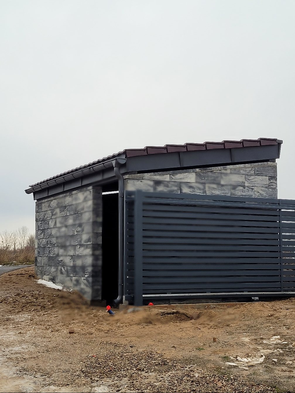 Nowoczesna wiata z szarej cegły z brązowym dachem i rynnami, przylegająca do szarego ogrodzenia lamelowego na tle pochmurnego nieba.