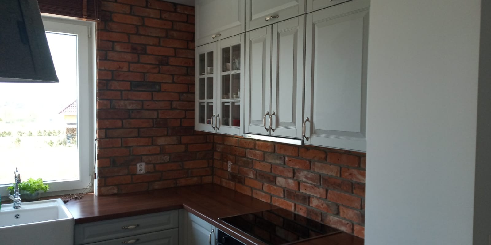 Kuchnia z ceglaną ścianą i białymi szafkami, widok na okno z rośliną w doniczce
