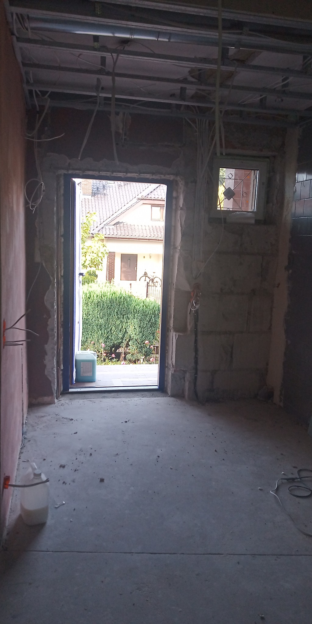 Wnętrze w trakcie remontu: surowe ściany, instalacje pod sufitem, widok na ogród przez otwarte drzwi balkonowe, butelka z płynem i kanister na podłodze.