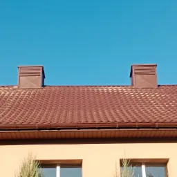 Czerwono-brązowy dach z blachodachówki z dwoma kominkami wentylacyjnymi na tle błękitnego nieba, widoczne rynny i fragment elewacji budynku.