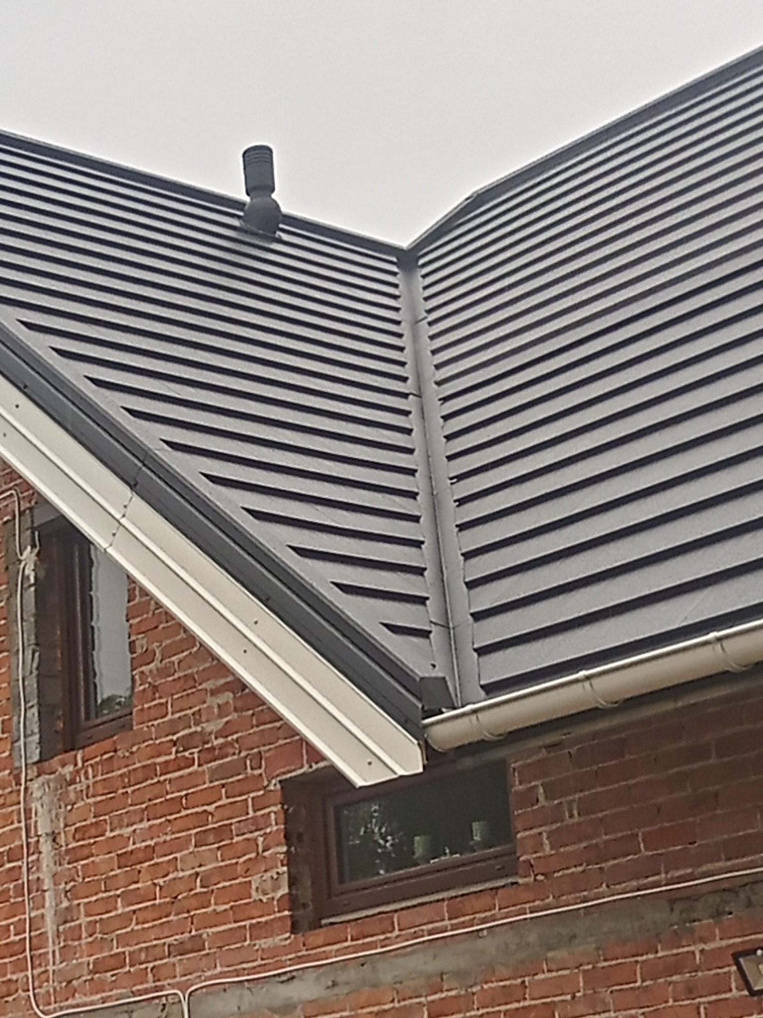 Ciemnoszara dachówka ceramiczna na domu z cegły, widoczny komin wentylacyjny i rynna z blachy powlekanej.