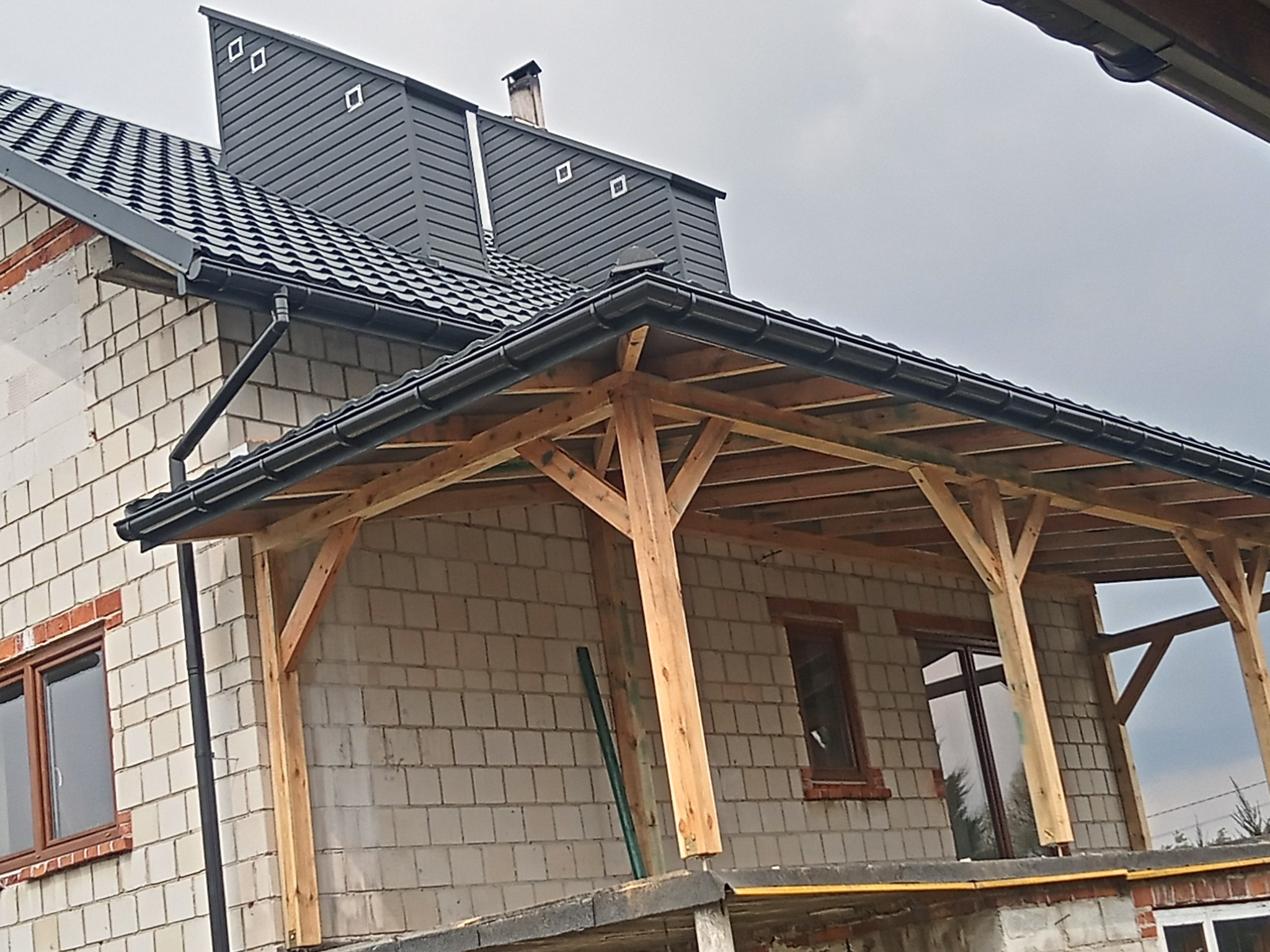 Nowoczesny dom w trakcie budowy z drewnianą konstrukcją zadaszenia tarasu, widoczne rynny i czarna dachówka oraz komin z szarą elewacją.