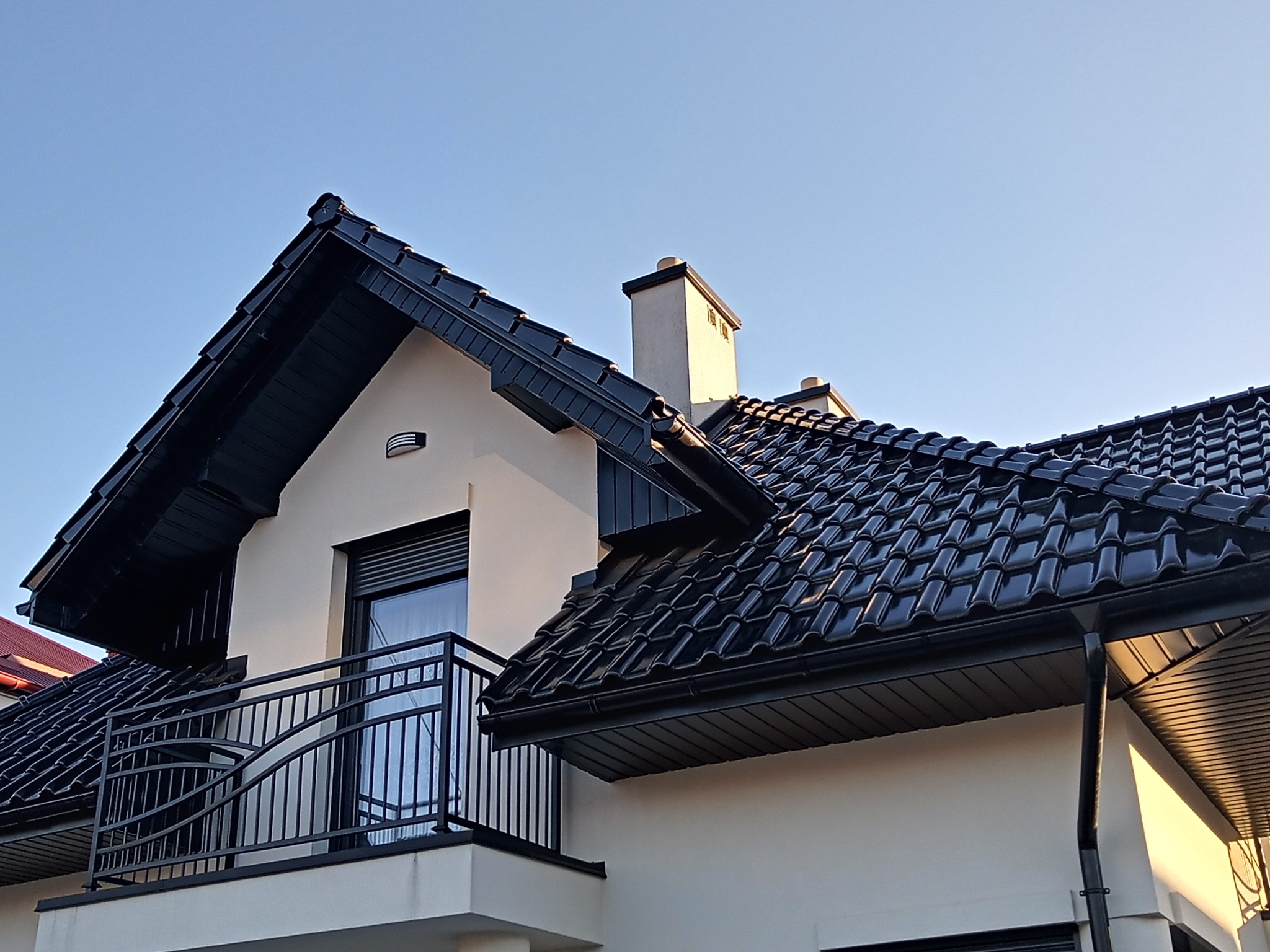 Nowoczesny dom z czarną dachówką ceramiczną, balkonem z metalową balustradą i widocznym kominem na tle błękitnego nieba.