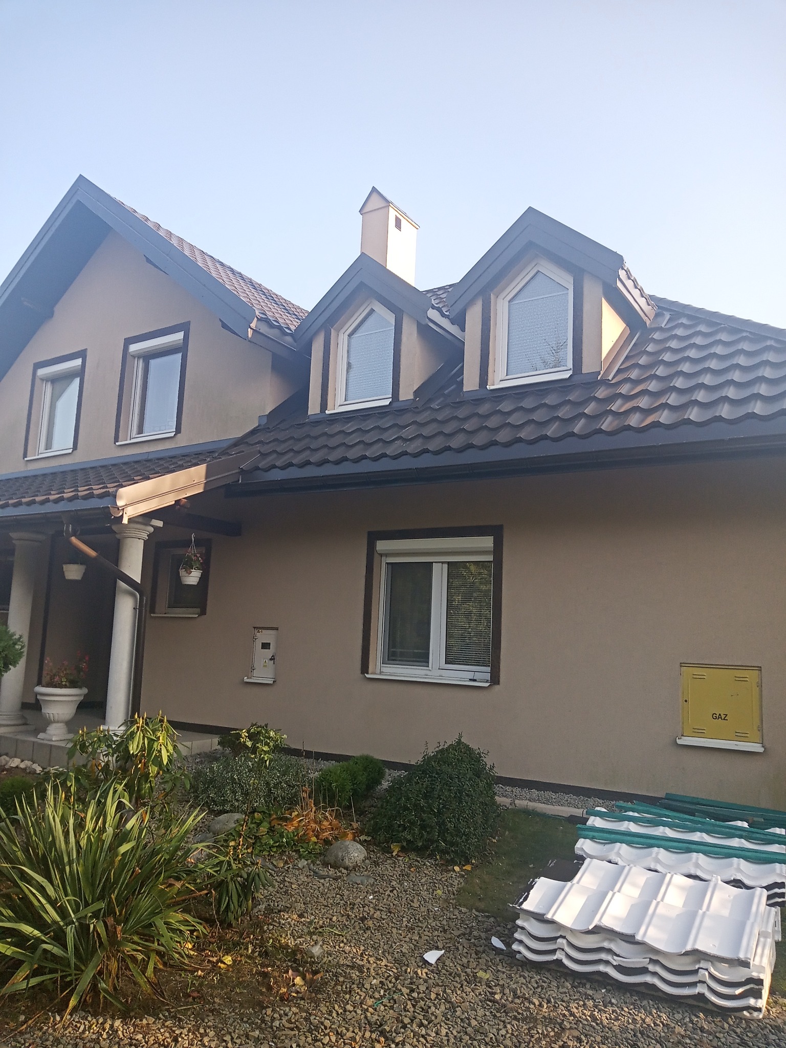 Dom jednorodzinny z nowym, brązowym dachem i stertą białych dachówek na ziemi przed budynkiem.