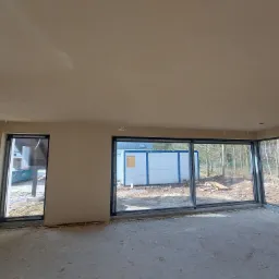 Wnętrze domu w stanie deweloperskim po nałożeniu tynków; widoczne duże okna z widokiem na las, surowe ściany i podłoga, wystające przewody elektryczne.