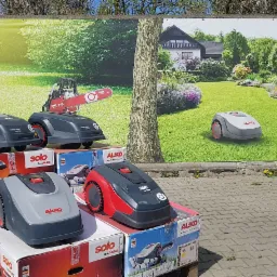 Ekspozycja robotów koszących marki AL-KO Solo na tle baneru reklamowego z motywem ogrodu i domu.