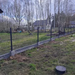 Nowoczesne ogrodzenie panelowe z podmurówką w kolorze szarym, widok na domy w tle i drzewa bez liści.